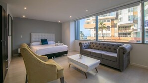 Une chambre d’hôtel moderne, équipée d’un lit, d’un canapé, d’un fauteuil et d’une table basse. Une grande fenêtre offre une vue sur un immeuble et des palmiers.