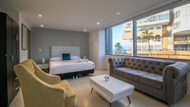 Une chambre d’hôtel moderne avec un lit, un canapé, un fauteuil et une table basse blanche.