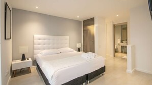 A modern bedroom with a large bed, bedside lamps, a nightstand, and a view into the bathroom. - Brand New 1 BR Apartment in Bocagrande (Cartagena)