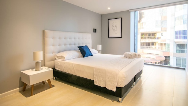 A modern bedroom with a large bed, a bedside table with a lamp, and a view of the city through large windows.
