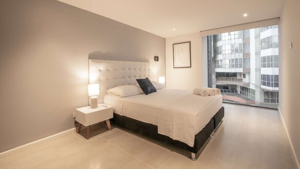 A modern bedroom with a large bed, a nightstand, and a view of a building through a large window. - 1 Bedroom Stunning Apartment in Bocagrande (Cartagena)