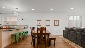 A modern kitchen with a wooden dining table and chairs, a bar area with stools, and a gray sofa in a living area.