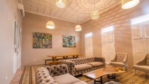A spacious room with a large sectional sofa, two armchairs, a wooden coffee table, and a dining table. There are three pendant lights hanging from the ceiling, and the walls are adorned with colorful artwork.