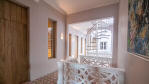 A hallway with patterned flooring, a wooden door, a white railing, and a view of a building with a spiral staircase.