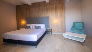 A modern bedroom with a large bed, a small round table, a teal armchair, and a white bench.