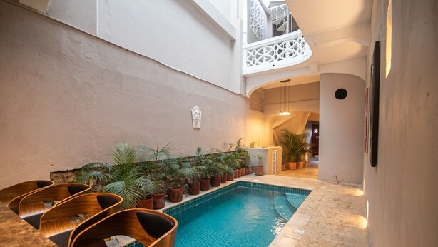 An indoor swimming pool with a tiled floor, surrounded by potted plants and wooden lounge chairs.
