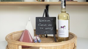 Een rieten dienblad met een fles wijn, een klein krijtbordje en een decoratief object.