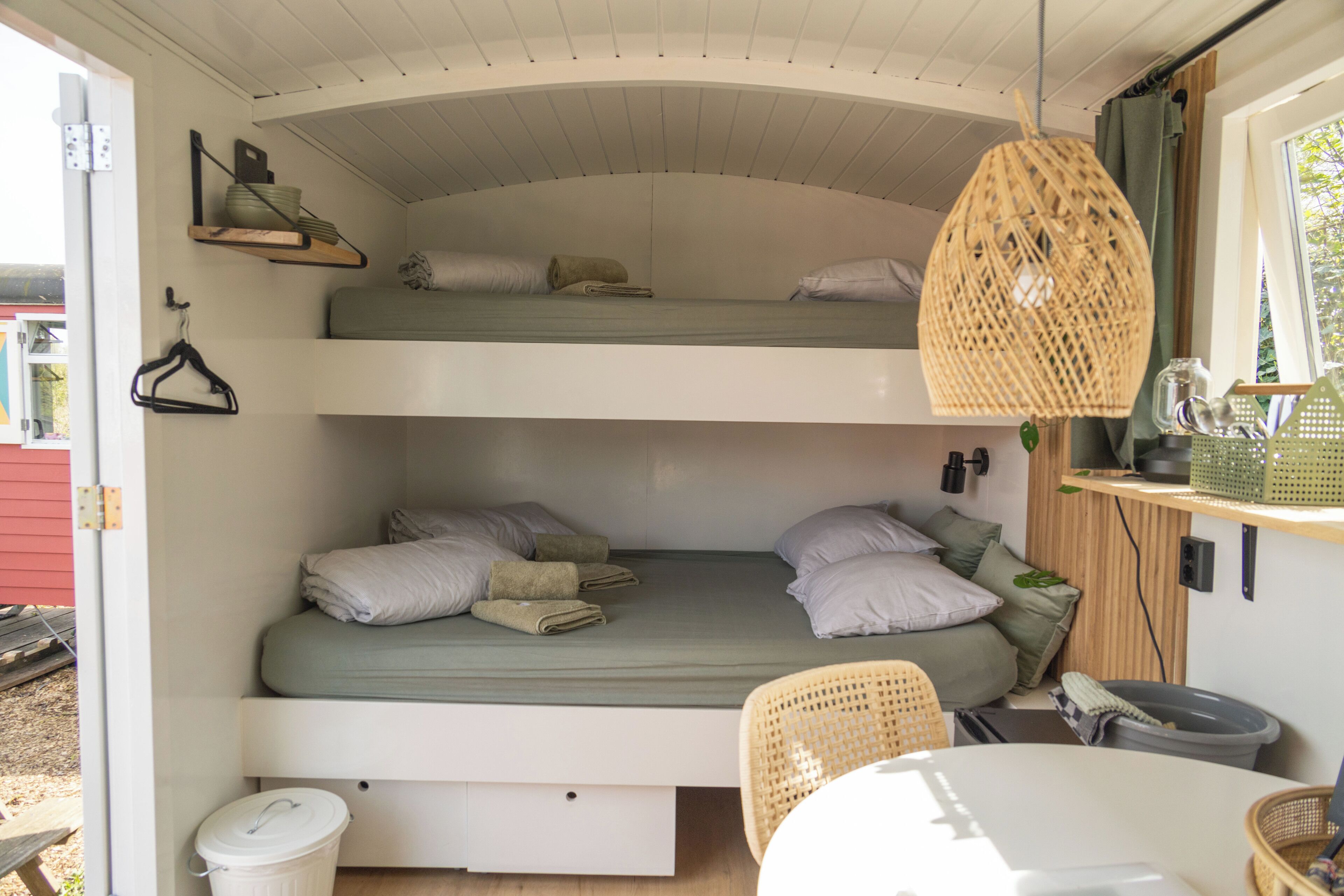 Quarto compacto com beliche de dois níveis, escrivaninha extensível, luminária pendente de vime e pequena mesa de jantar.