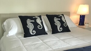 Un lit avec une literie blanche et bleu marine, deux coussins bleu marine ornés de motifs représentant des hippocampes, une enseigne bleue accrochée au mur avec l’inscription « Bienvenue à Vero Beach », et une lampe de chevet.