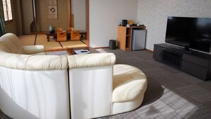 A modern living room with a sectional sofa, a flat-screen TV, and a small cabinet. - Inn Juraku Formerly Spa Riraku (Toon)