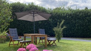 An outdoor seating area with a wooden table and chairs, a large umbrella, and a hedge in the background.