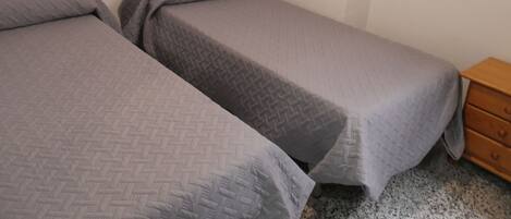 Two single beds with grey bedding, a wooden headboard, a wooden nightstand, and a small round mirror on the wall.