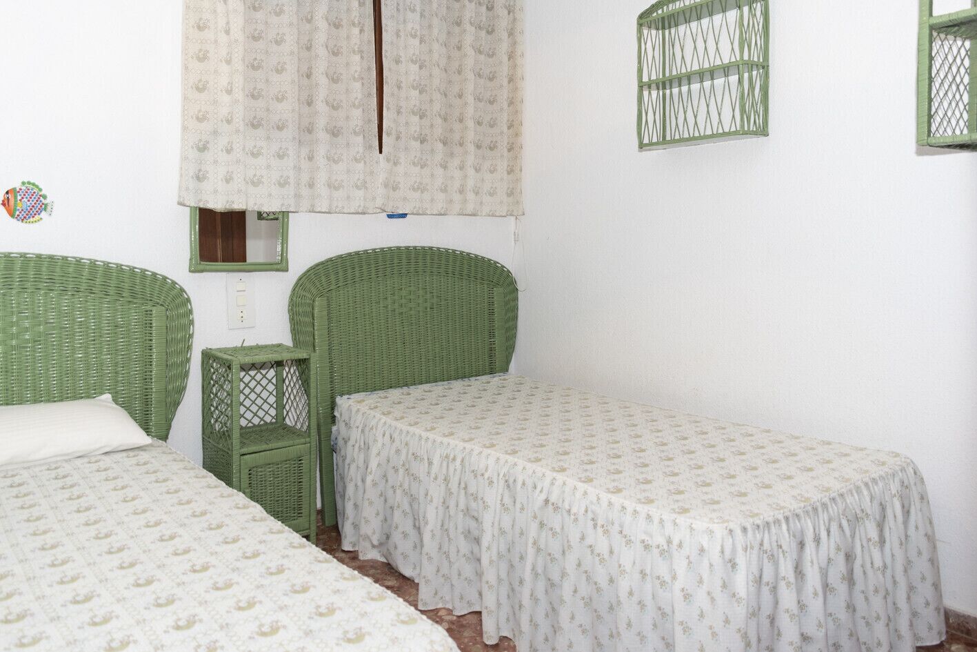 Een kamer met twee groene rieten bedden, een klein groen bijzettafeltje en een raam met gordijn.