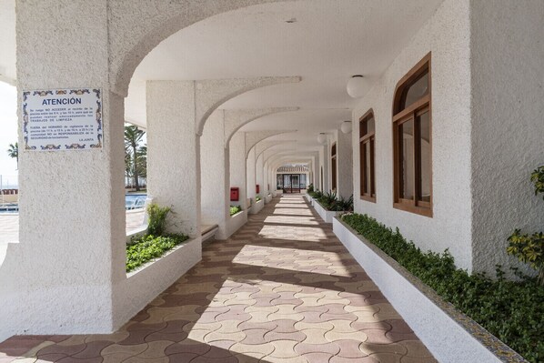 A covered walkway with white walls and wooden-framed windows, leading to a building entrance.