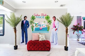 A modern hotel lobby with a red tufted sofa, two potted palm trees, and a wall with a poster of a man in a suit and another in a pink shirt. - 80’s Paradise golf cart, rooftop deck heated pool (Indian Rocks Beach)