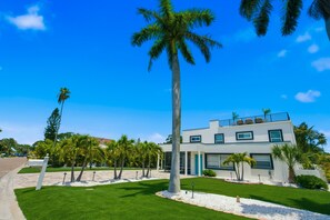 A modern two-story building with a flat roof, surrounded by a well-maintained lawn and palm trees. - 80’s Paradise golf cart, rooftop deck heated pool (Indian Rocks Beach)
