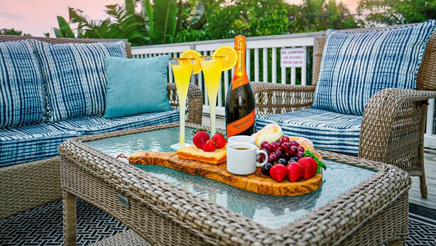 A wicker outdoor seating area with a glass table set with a bottle, glasses, fruit, and a sign indicating no jumping or driving from the deck.