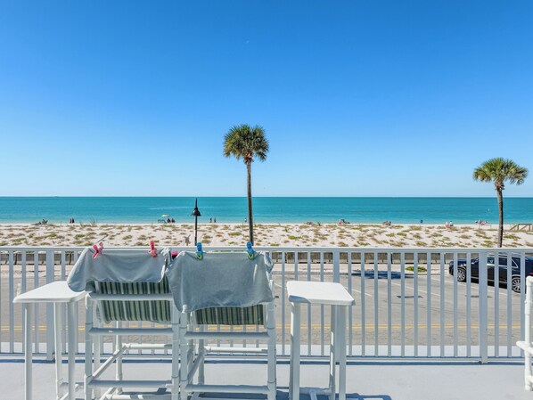 A beach view with a white railing, two chairs, and a table with a cloth.