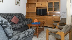 Una sala de estar con un sofá, una mesa de centro y un televisor. Se observan armarios de madera y una silla.