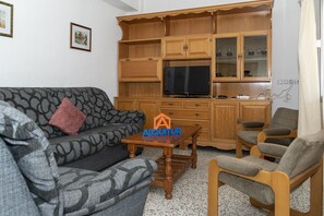A living room with a sofa, a coffee table, and a TV. There are wooden cabinets and a chair. - MIRALMAR, 10º-69 (Cullera)