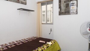 Una habitación pequeña con cama individual, ventana, repisa en la pared y ventilador de pared.