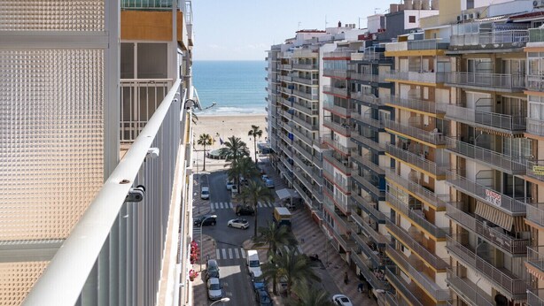 Un edificio de gran altura con varios balcones que dan a una playa y una calle con coches y peatones.