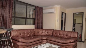Ein braunes, ledernes Ecksofa, ein Metallhocker und ein kleiner Tisch mit Papieren in einem Wohnzimmer mit großem Fenster und Wandklimaanlage.