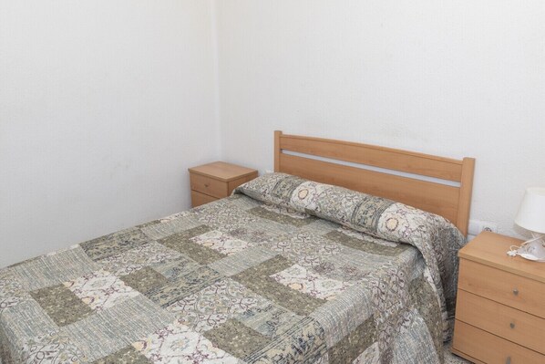A single bed with a patterned quilt, a wooden headboard, a nightstand with a lamp, and a small drawer unit. - ESTIRPE, 2º-3 (Cullera)
