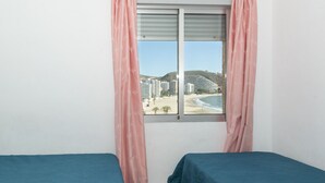 Een kamer met twee bedden, een raam met roze gordijnen en uitzicht op een strand en gebouwen.