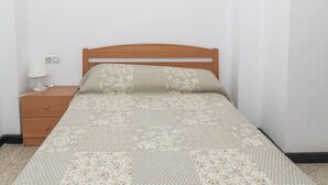 A single bed with a floral-patterned bedspread and pillows, a wooden nightstand with a lamp, and a power outlet on the wall. - ESTIRPE, 2º-4 (Cullera)