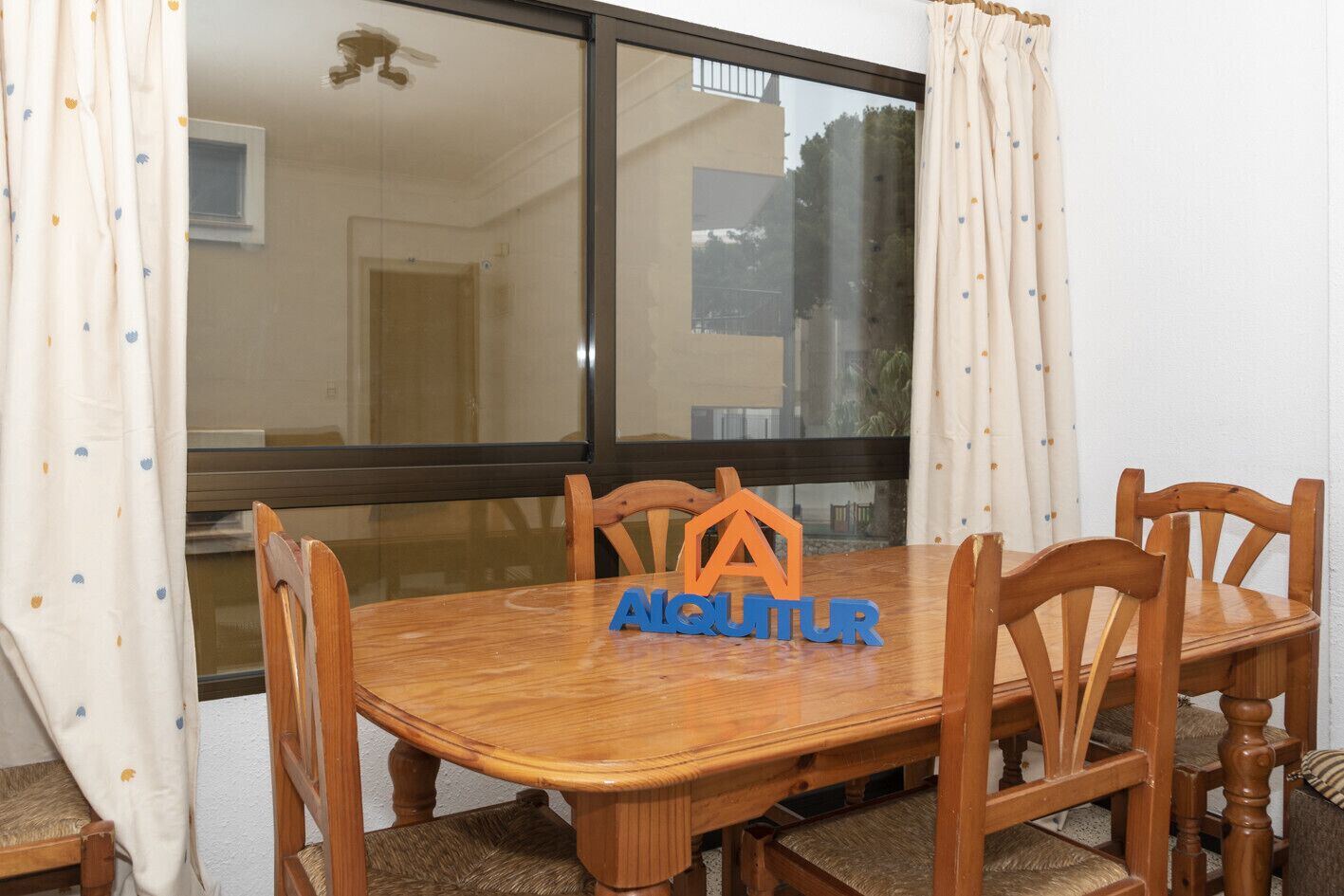 Een eetgedeelte met een houten tafel en stoelen, een raam met gordijnen, en een logo op de tafel.