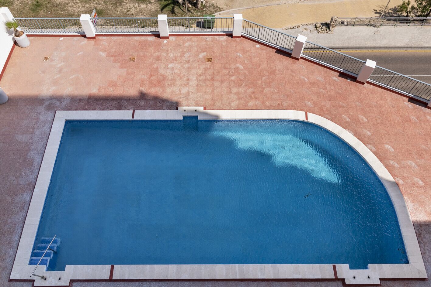 A rectangular swimming pool with a white border, surrounded by a red-tiled deck and a metal railing.