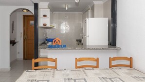 A kitchen with a white refrigerator, a sink, and a countertop. There are two wooden chairs and a table with a newspaper-patterned tablecloth. - DOSEL, 6º-30 PARKING 30 (Faro de Cullera)