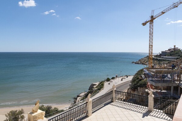 A coastal view with a construction crane, a beach, and a building with a balcony overlooking the sea.
