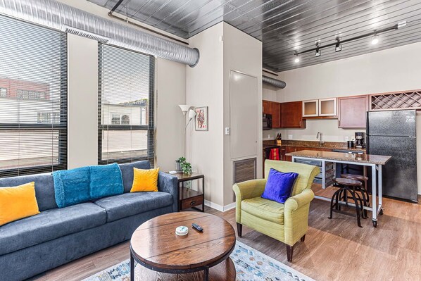 A modern living room with a gray sofa, yellow and blue cushions, a round wooden coffee table, and a green armchair. A kitchen area with wooden cabinets and a stainless steel refrigerator is visible in the background.
