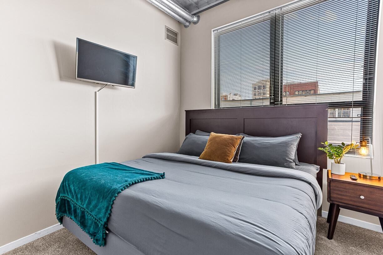 Ein Schlafzimmer mit einem Bett, einem an der Wand montierten Fernseher, einem Fenster mit Jalousien und einem Nachttisch mit einer Pflanze.