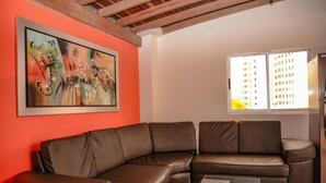 A living room with a brown leather sofa, a painting on the wall, and a window with a view of buildings.