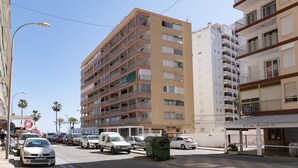 Un edificio de varios pisos con balcones, una calle con coches estacionados y un cielo despejado.