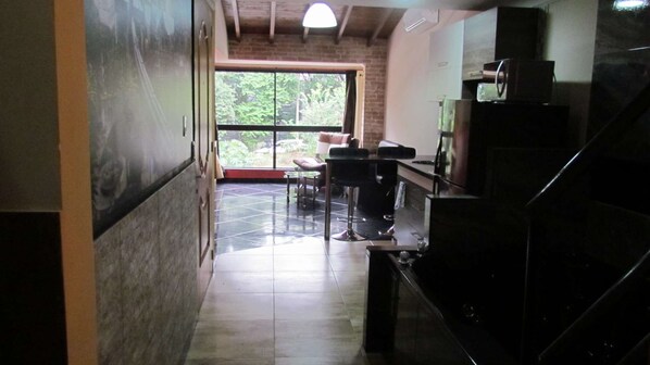 A hallway with a tiled floor, a seating area with chairs and a table, a large window with a view of greenery, and a staircase with a black railing.