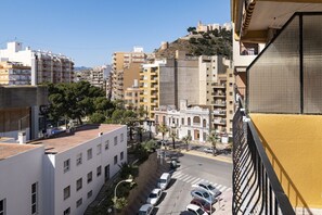 A cityscape with a mix of residential and commercial buildings, a street with parked cars, and a hill with historical structures in the background. - MAR Y MONTAÑA 7º-119 (Cullera)