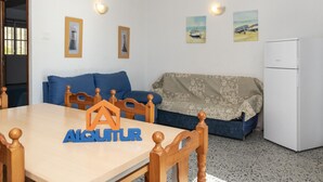 A room with a wooden dining table, blue sofa, and a patterned blanket. There are framed pictures on the wall and a white refrigerator. - TRIANON, 5º-29 (Cullera)