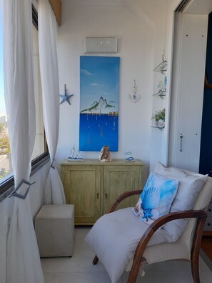 Une pièce avec vue sur la mer, un tableau représentant un paysage côtier, un meuble en bois orné d’objets décoratifs et un fauteuil avec un coussin.