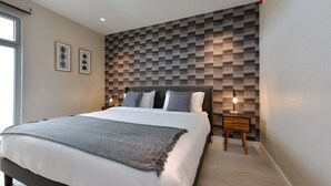 A modern bedroom with a geometric-patterned headboard, a large bed with white linens, a grey throw blanket, and a bedside table with a lamp. - Brand New Apartment, Panoramic Ocean Views (Noord)