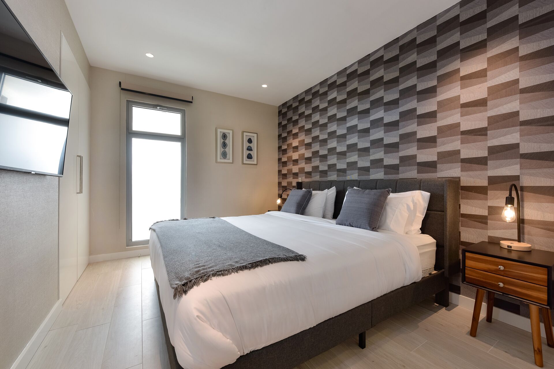 A modern bedroom with a large bed, a geometric-patterned headboard, a bedside table with a lamp, and a window with blinds.
