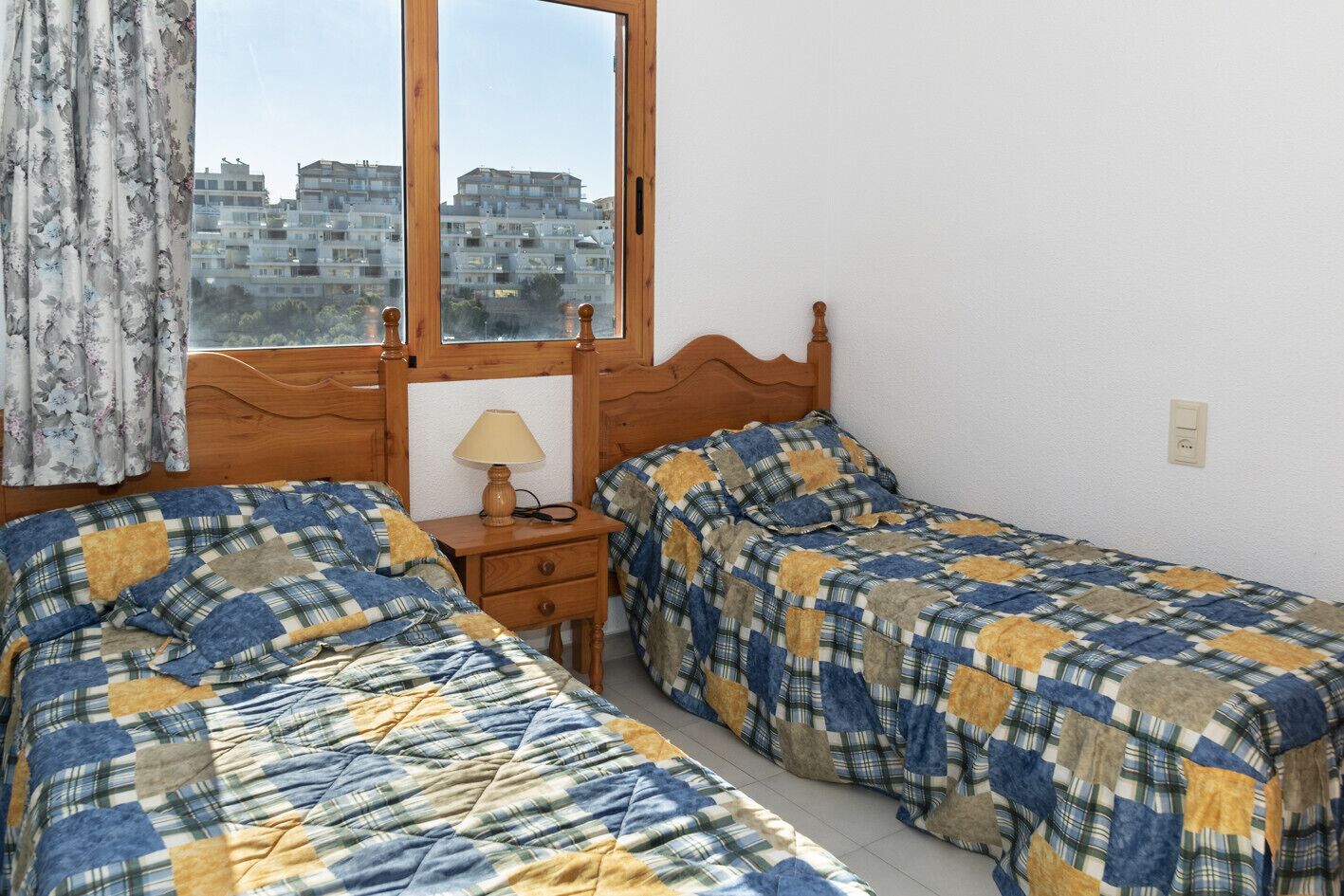 A bedroom with two beds, a wooden headboard, a nightstand with a lamp, and a window with a view of buildings.