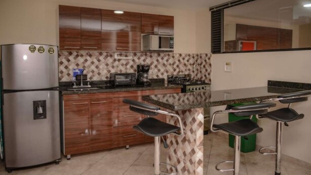 Una cocina moderna con armarios de madera, un refrigerador de acero inoxidable, un microondas, una cafetera y una estufa a gas. Hay dos taburetes de bar en una pequeña isla.