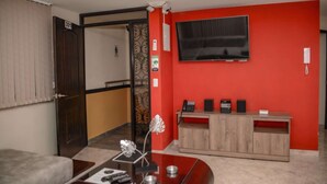 Habitación de hotel con una pared roja, un televisor sobre un mueble de madera y una puerta que da a otra habitación.