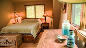 A cozy bedroom with a bed, bedside lamps, a wooden nightstand, a wicker chest, a wooden table with a bowl and a vase, and a window with blinds. - Luxury Lakefront: Hot Tub, Fireplace, Pet Friendly, Arcade, Watercraft, + more! (Lake Ariel)