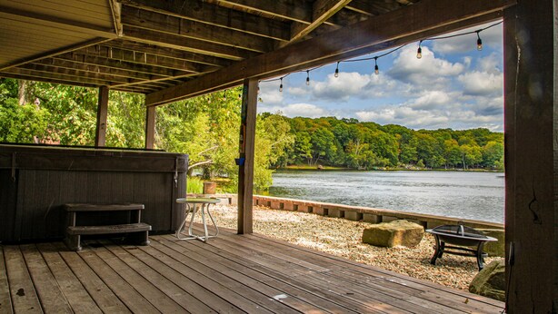 A wooden deck with a bench and a table, overlooking a lake with trees and a cloudy sky. - Luxury Lakefront: Hot Tub, Fireplace, Pet Friendly, Arcade, Watercraft, + more! (Lake Ariel)