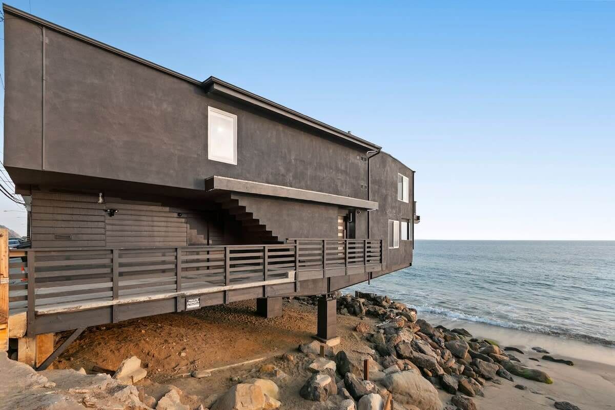 A modern coastal house with a balcony, built on a rocky shoreline.